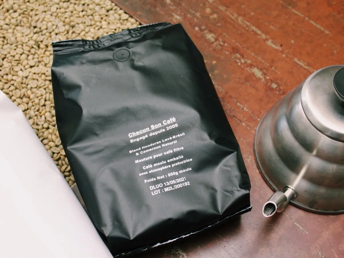 Sachet de café moulu Chacun Son Café posé sur des grains de café vert arabica, avec bouilloire